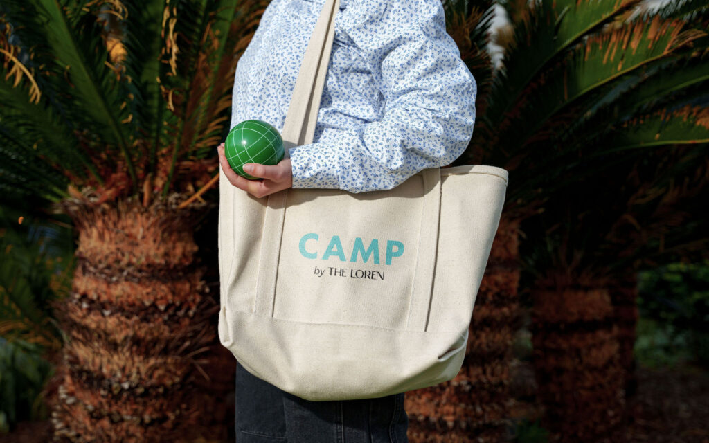 Person holding a green ball and wearing a "CAMP by THE LOREN" tote bag, standing in front of palm trees.