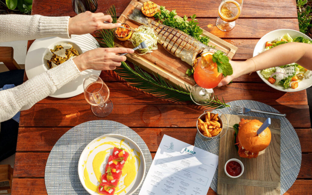 Overhead view of a table with grilled fish, burger, salad, fries, drinks, and two people reaching for food.