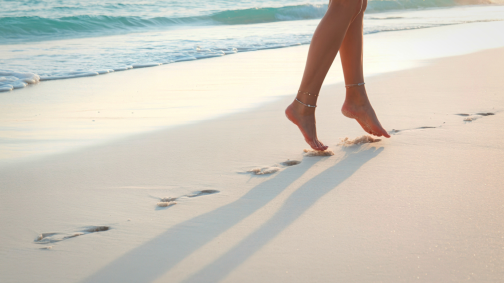 A woman walking along the coastline with blue ocean waves in the background, leaving footprints in the sand.