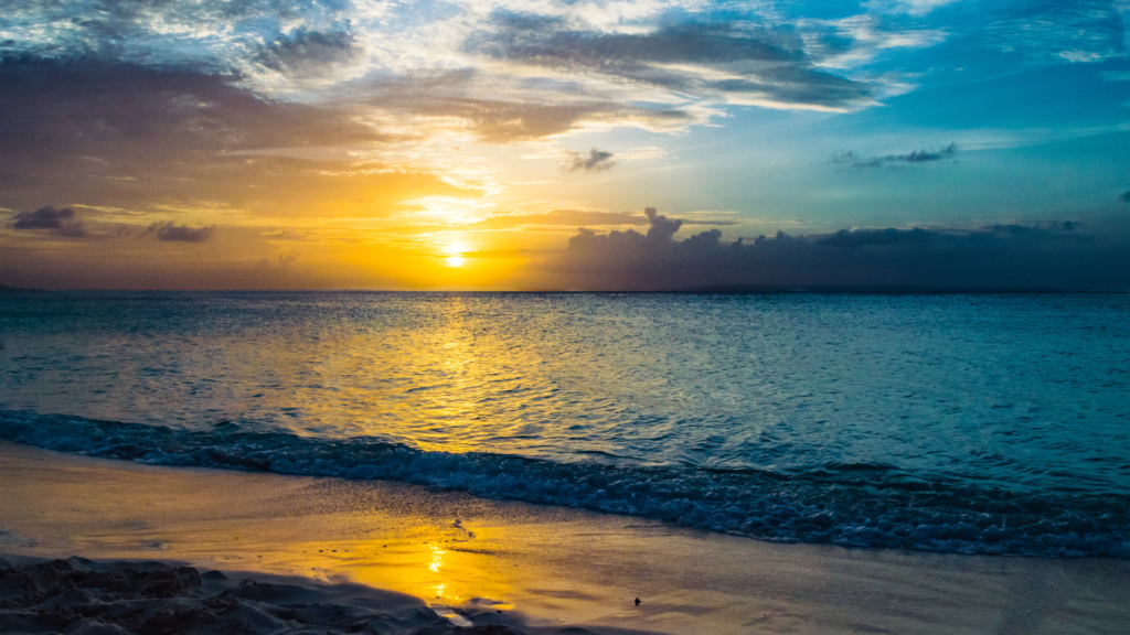 A shoreside view of the ocean at sunset, with the yellow light of the fading sun reflecting off of calm blue waters.
