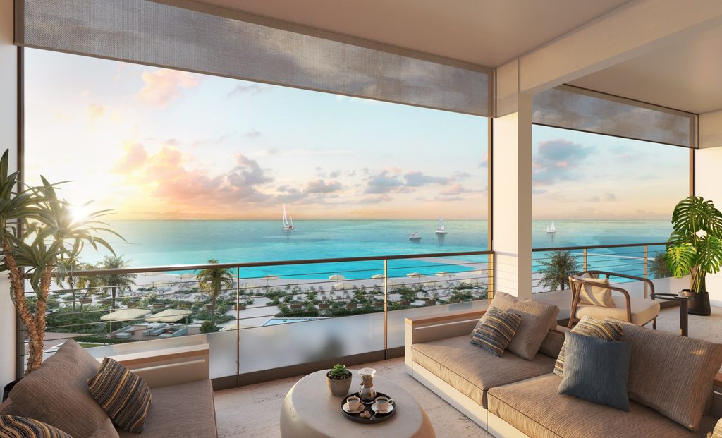 Modern beachfront balcony with lounge seating overlooks ocean, palm trees, and sailboats at sunset.