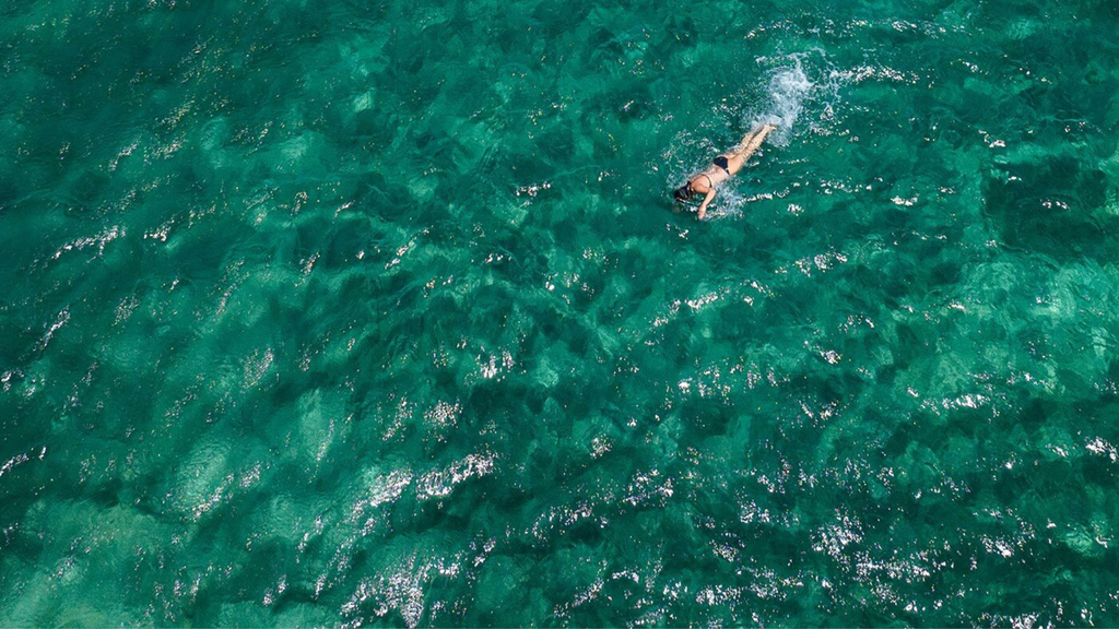 A person swims alone in clear turquoise water, viewed from above on a sunny day.